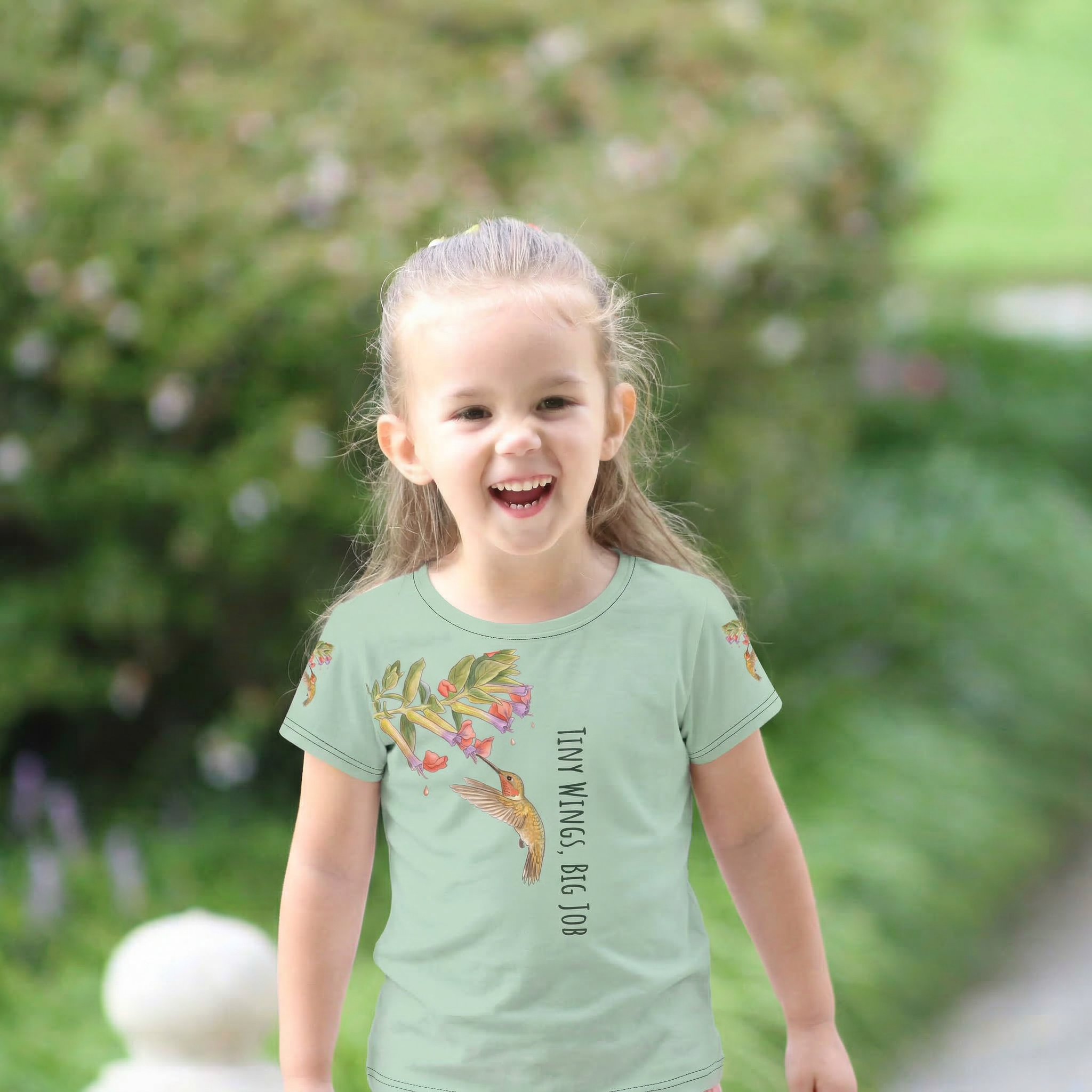 "Tiny Wings Big Job" all-over print toddler t-shirt featuring hummingbirds and vertical text on a whimsical floral background.
