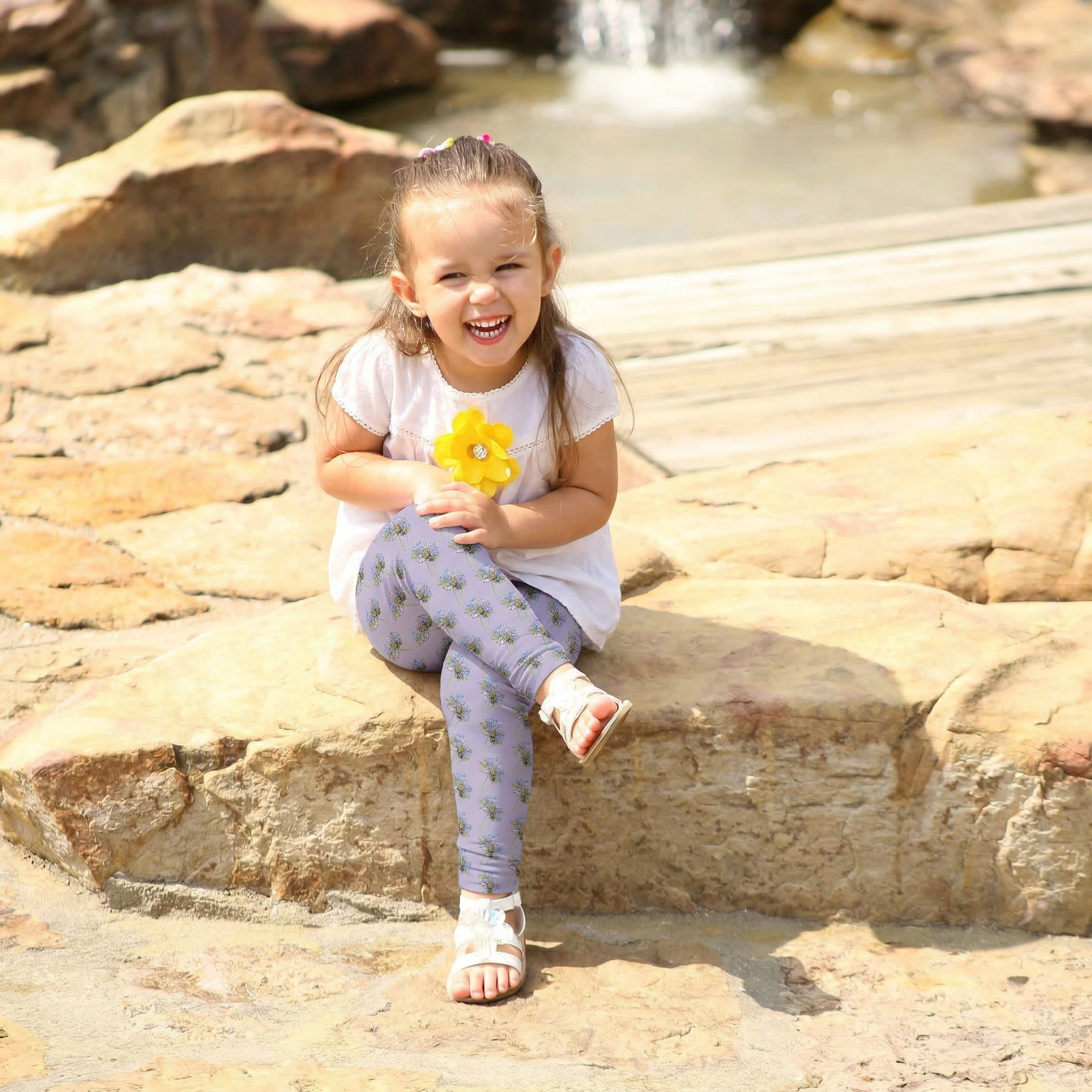 A young girl sitting on rock near waterfall wearing dark lavendar leggings with a vibrant hummingbird and floral nectar quest print.