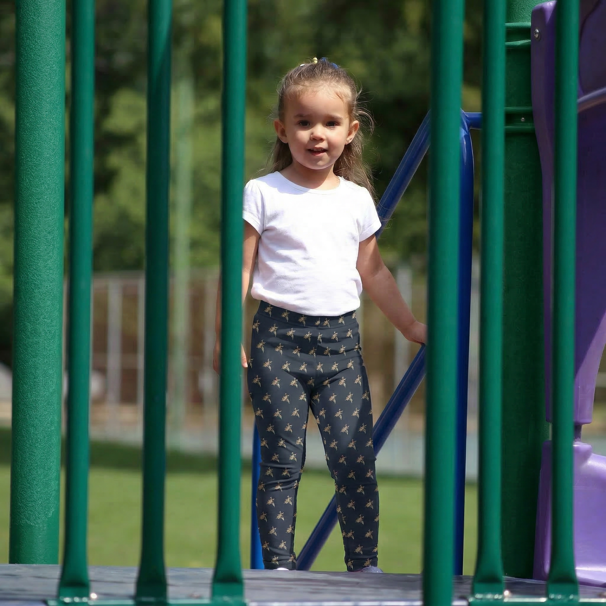 A young girl playing on a green playground slide wearing dark navy leggings with a vibrant hummingbird and floral nectar quest print.