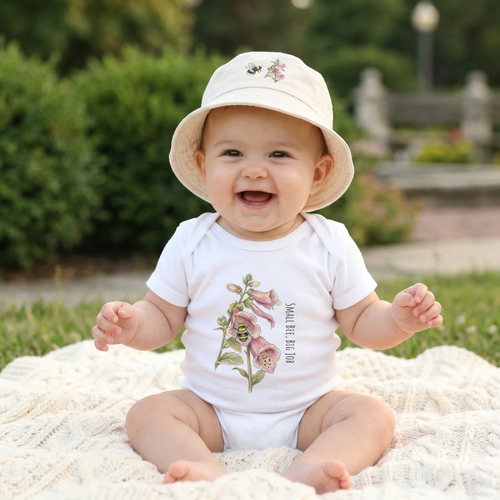 Baby girl sitting on a blanket wearing a white 100% cotton baby one-piece with 'Small Bee, Big Job' bumblebee and foxglove illustration on the front.