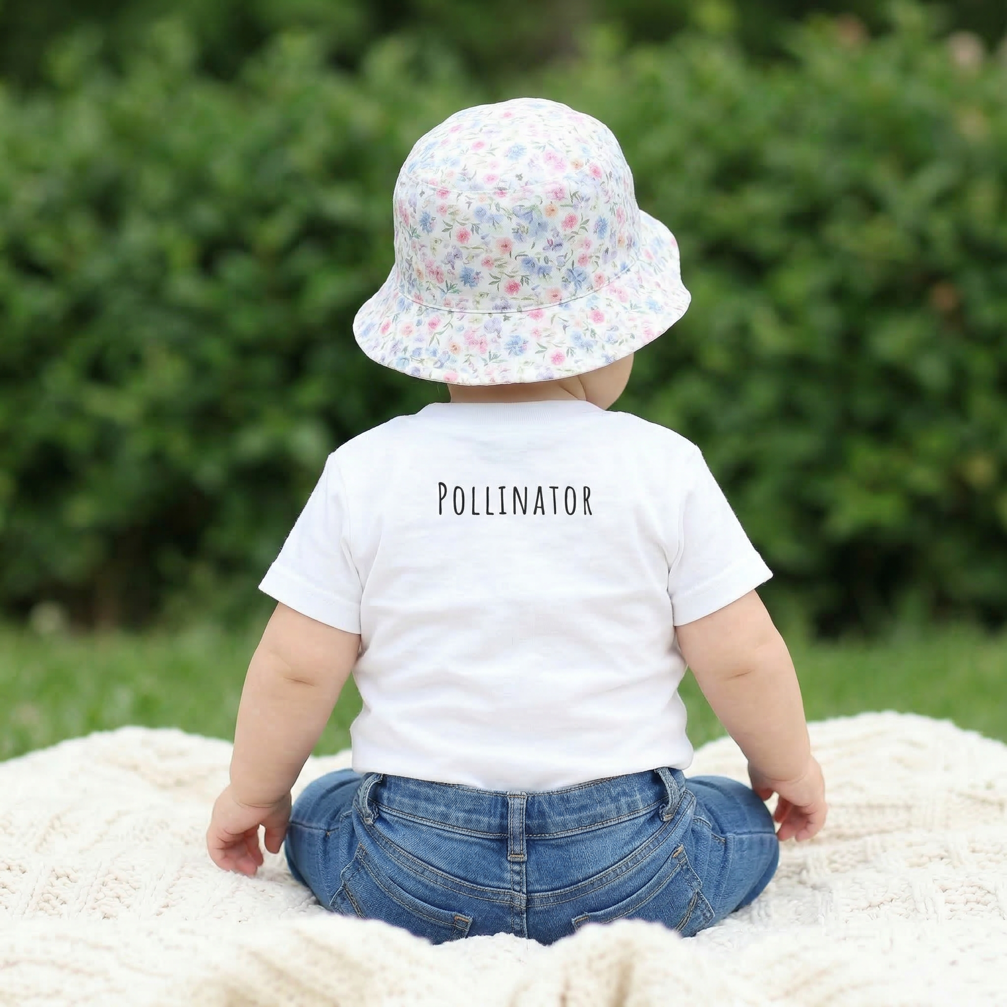 Child wearing a white shirt with 'POLLLINATOR' text, sitting on a blanket outdoors.