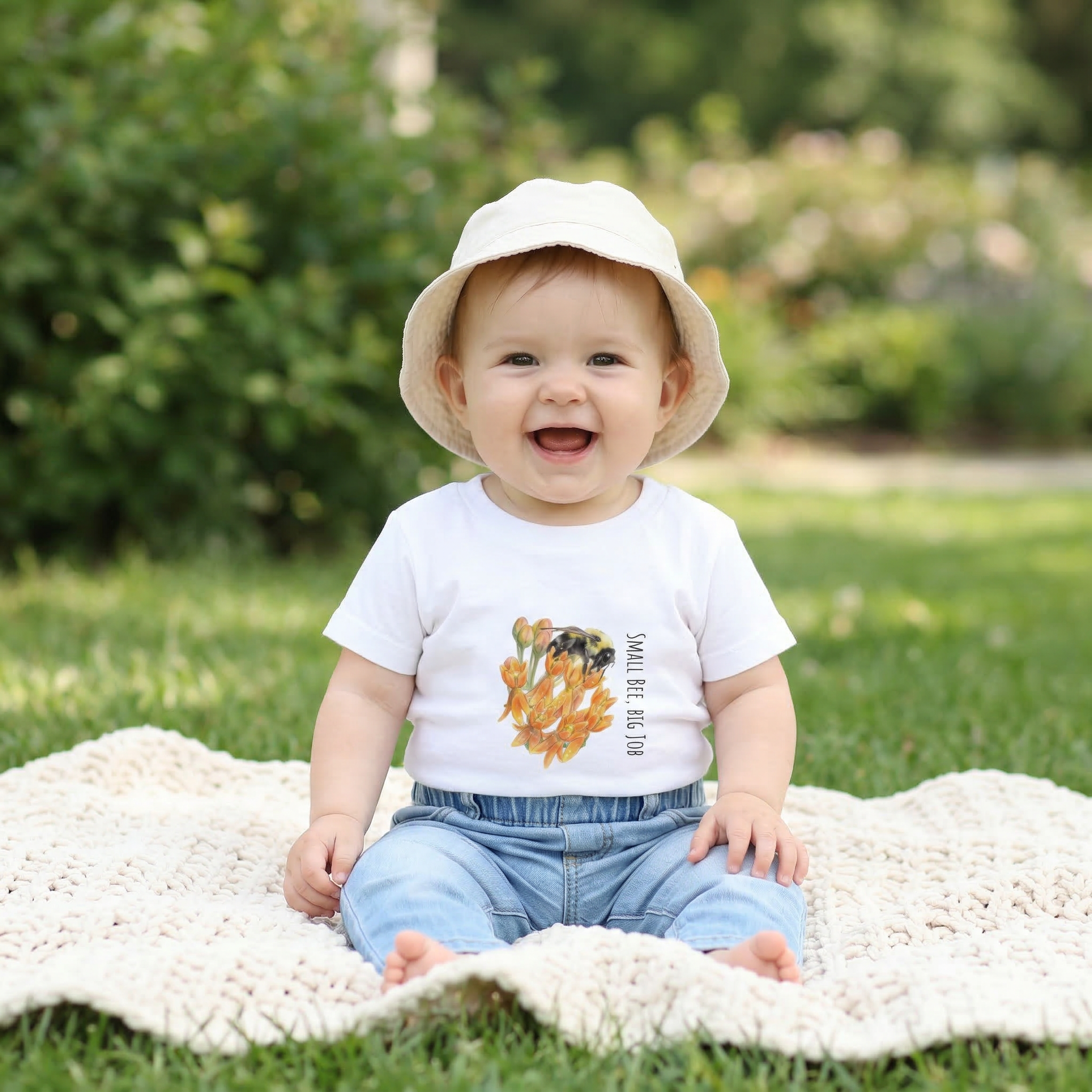Baby sitting on a blanket outdoors wearing a white shirt with a graphic bee and butterfly weed design and a beige hat.