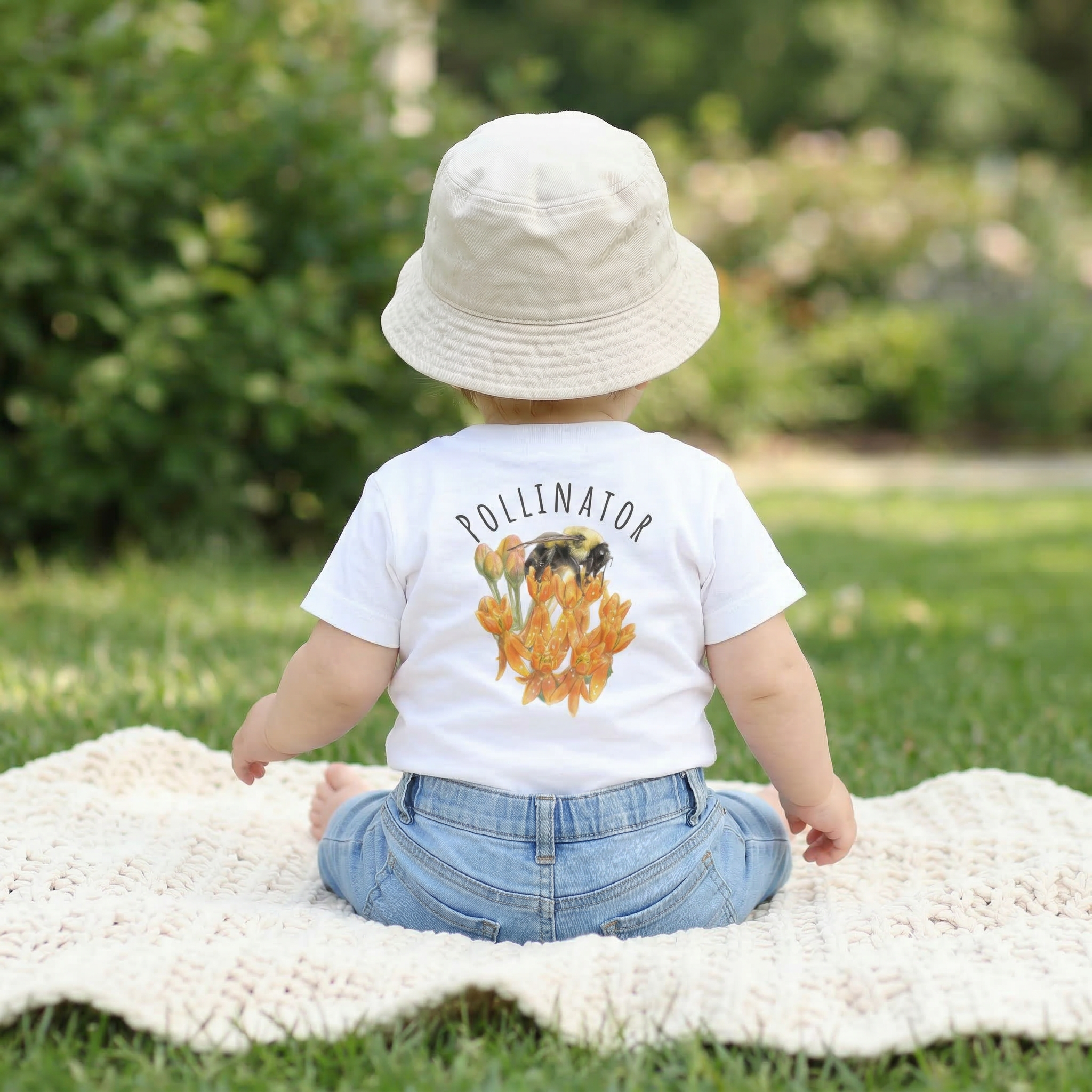 back view of a Child wearing a 'Pollinator' t-shirt with a graphic of bees and flowers, sitting on a blanket in a garden.