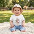 Baby sitting on a blanket outdoors wearing a white shirt with floral design and text "small bee, big job" and blue jeans.