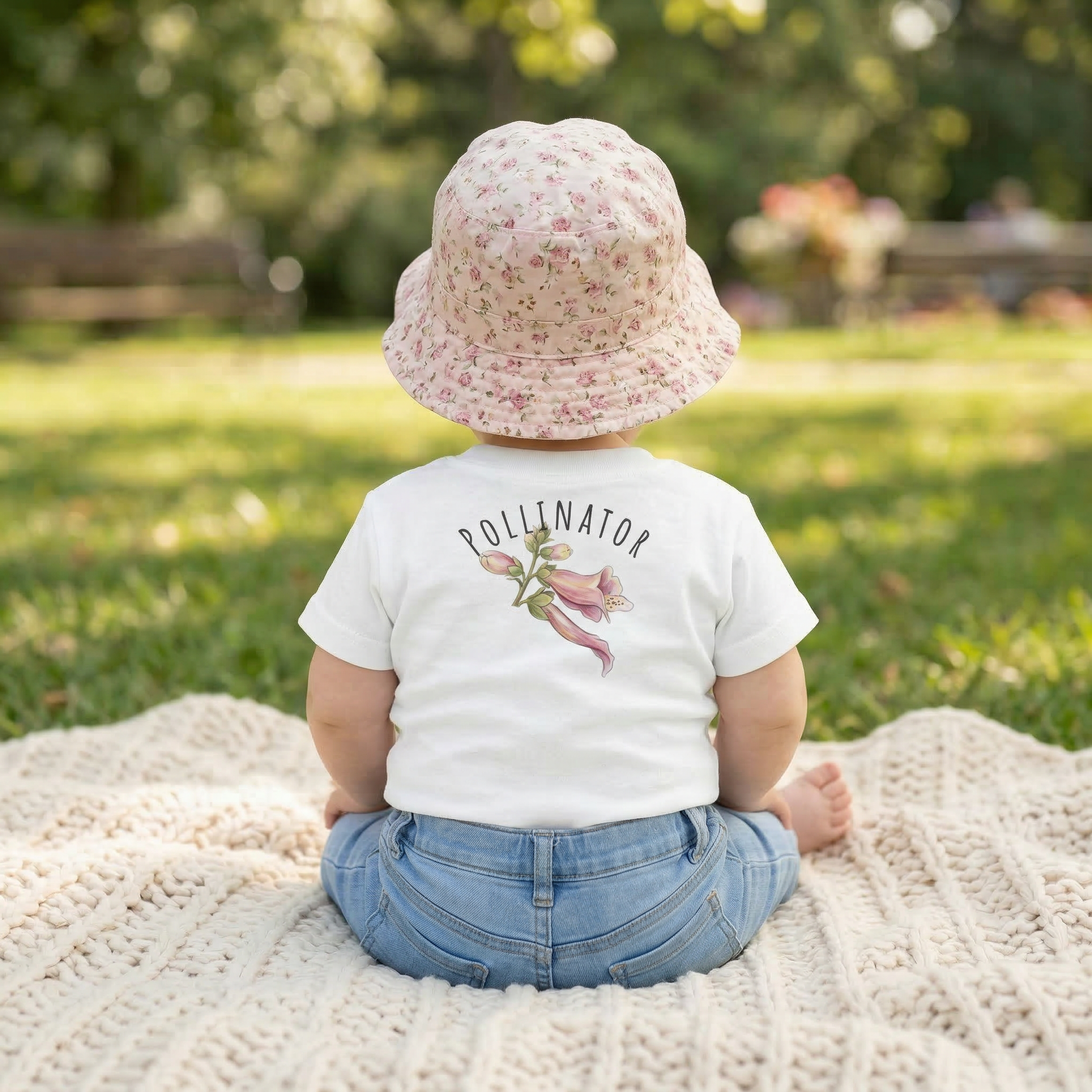 Child wearing a white t-shirt with 'Pollinator' text and flower graphic, sitting on a blanket outdoors.