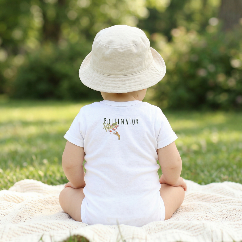Child wearing a white 'Pollinator' shirt and hat, sitting on a blanket in a grassy area.