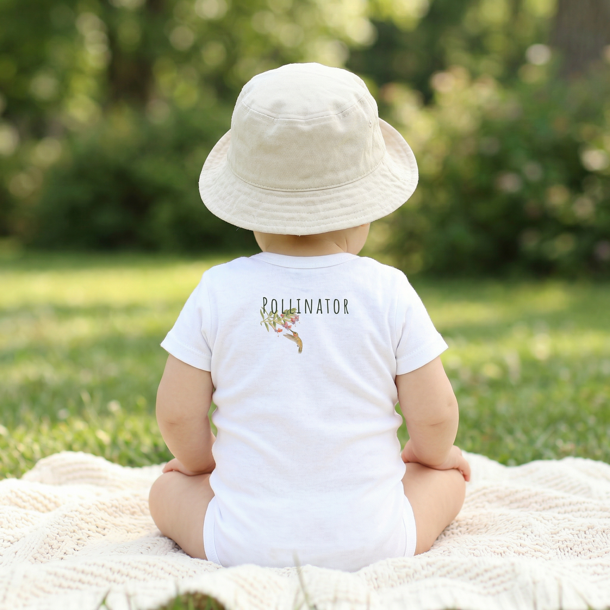 Child wearing a white 'Pollinator' shirt and hat, sitting on a blanket in a grassy area.