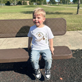 Child sitting on a park bench wearing a white t-shirt with hummingbird graphics and text "tiny explorer"