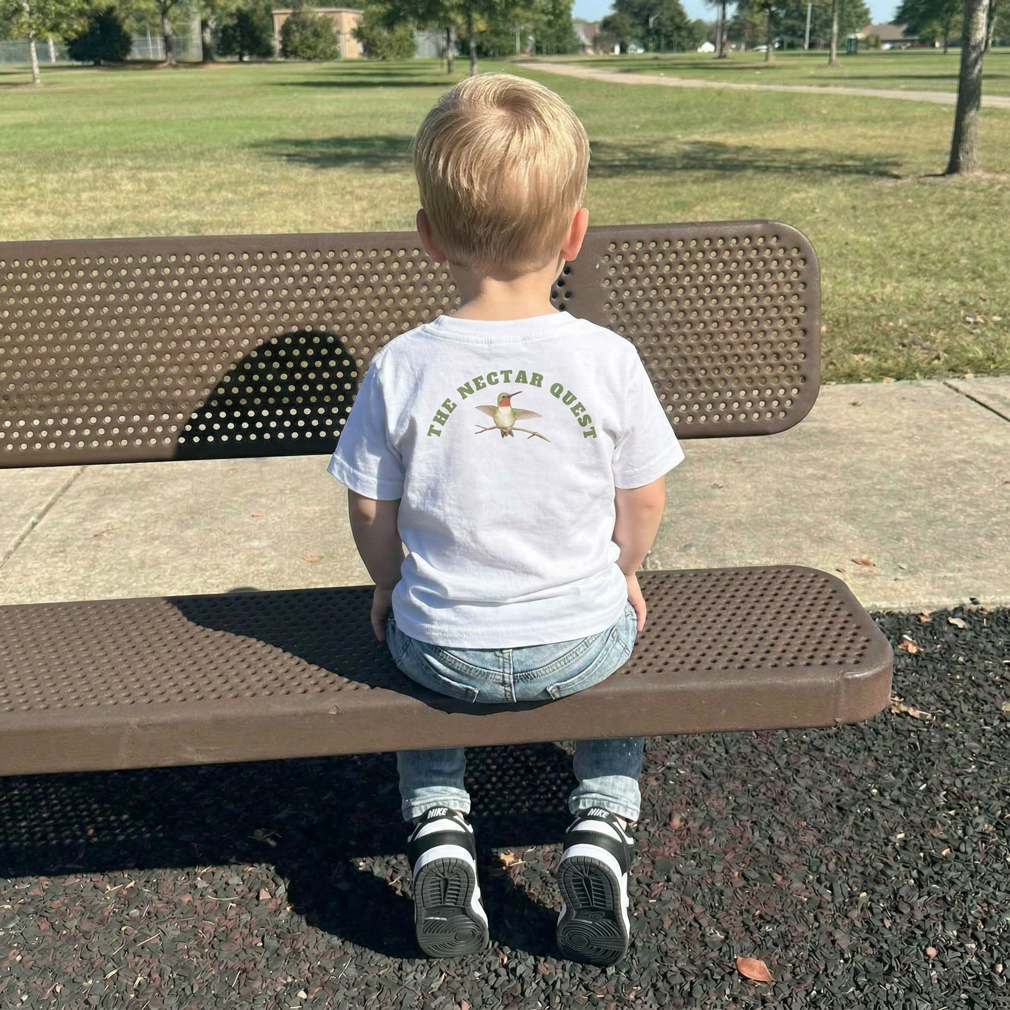 Back view of a Child sitting on a bench in a park wearing a white t-shirt with text "The Nectar Quest" and hummingbird graphics. 