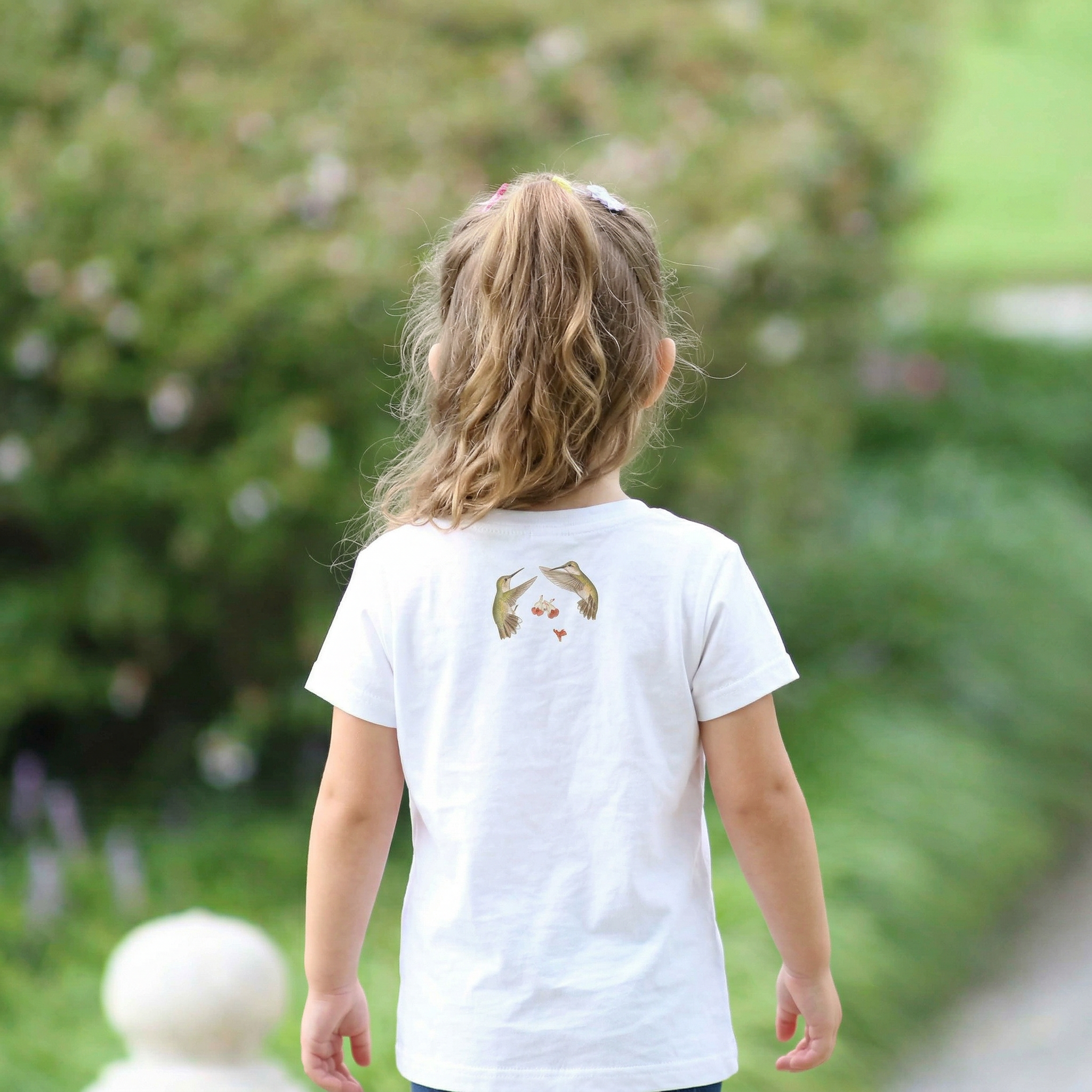 Back view of Child wearing a white t-shirt with hummingbird graphics outdoors.