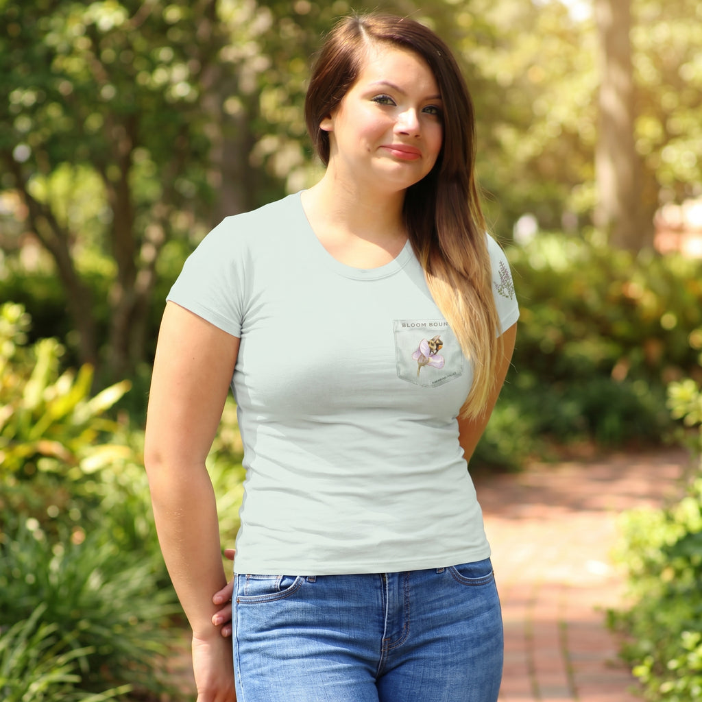 Woman wearing a light sage t-shirt with a bee in a flower on a faux pocket desigh in an outdoor setting