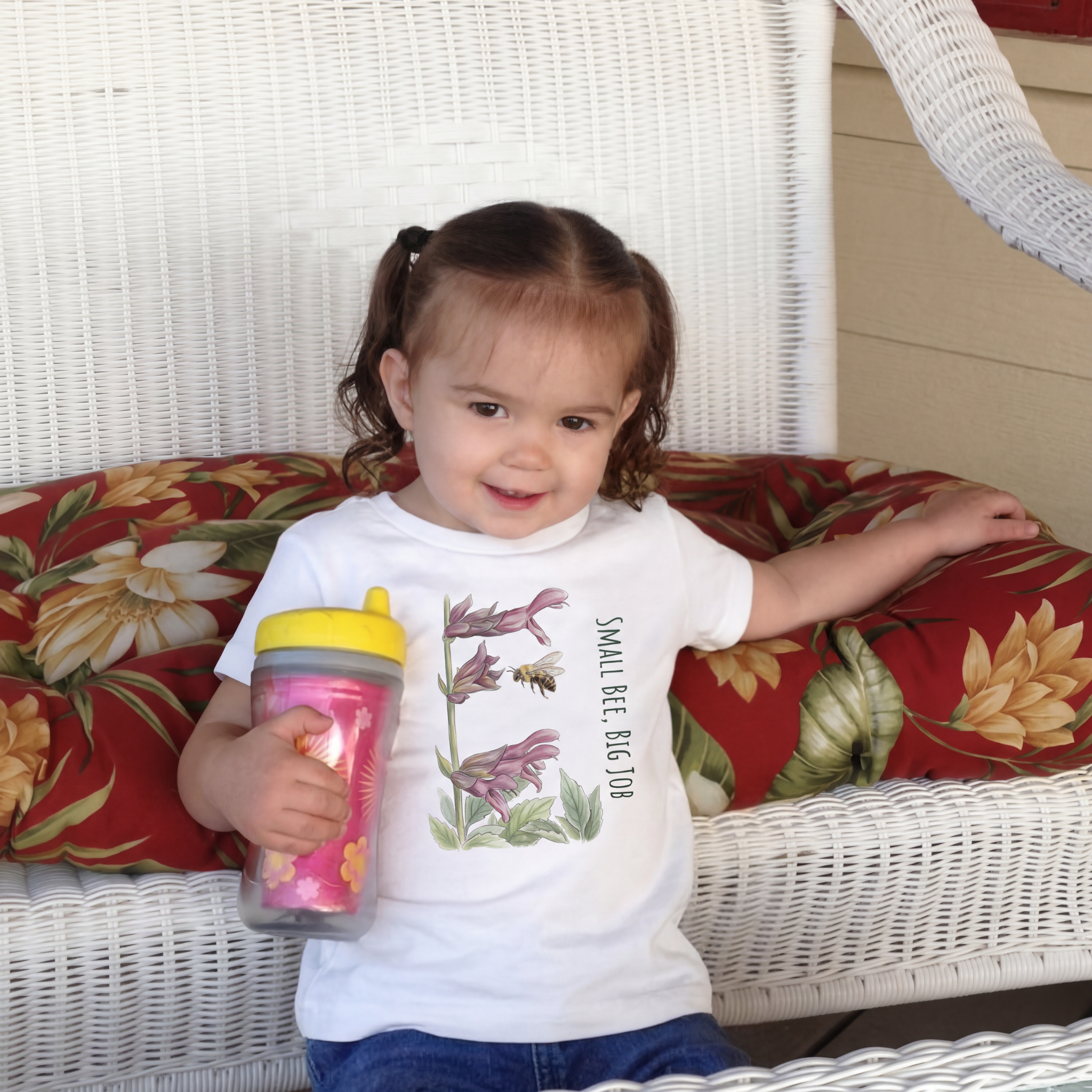 Child holding a sippy cup on a floral cushioned chair, wearing a Small Bee, Big Job classic cotton Tee