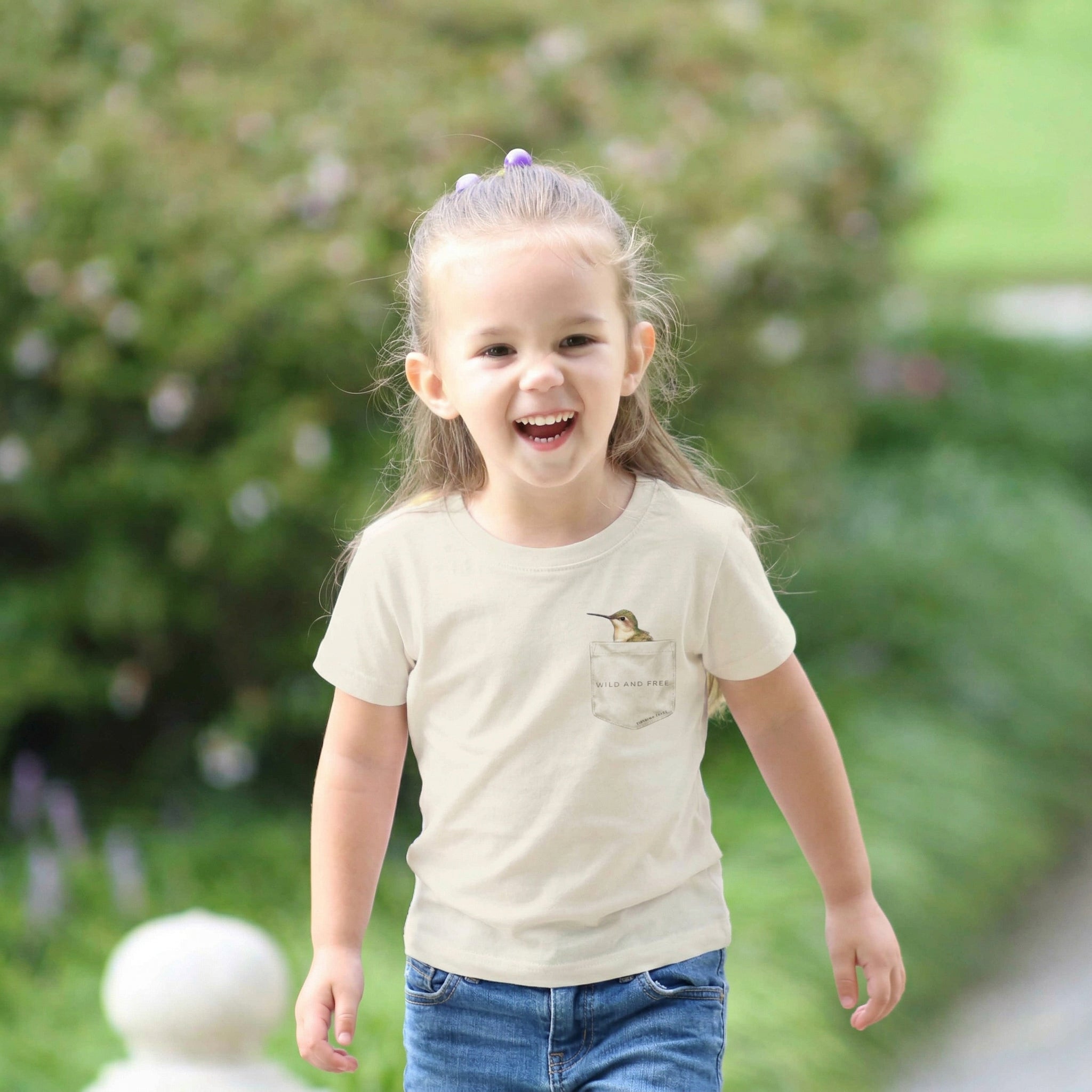 toddler wearing a cream colored faux pocket tee with hummingbird design. Tinying Tales