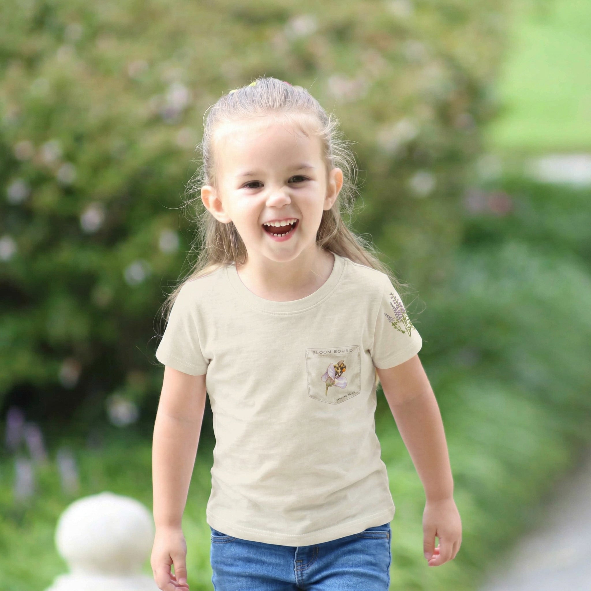 Child wearing a beige t-shirt with a small design, standing outdoors with greenery in the background. Tinywing Tales