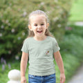 Child wearing a light sage t-shirt with a small design on a faux pocket design of a bee and flower. Text "Bloom Bound" Tinywing Tales