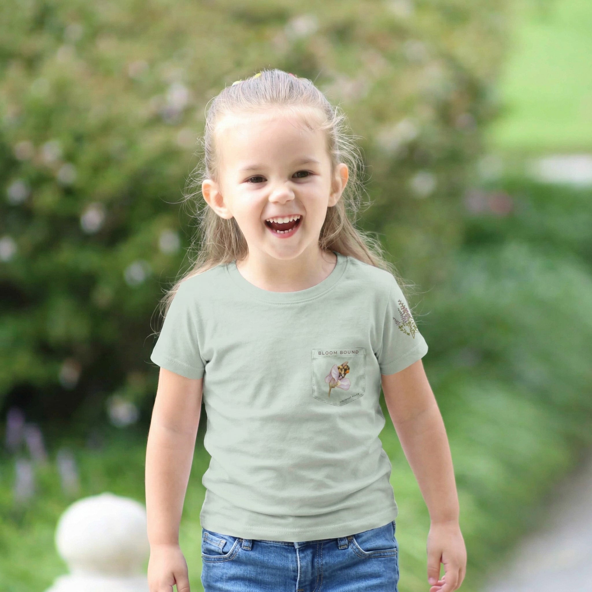Child wearing a light sage t-shirt with a small design on a faux pocket design of a bee and flower. Text "Bloom Bound" Tinywing Tales